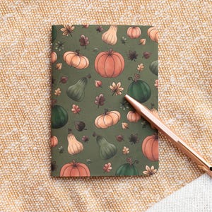 Może przedstawiać: Zielony notes z wzorem pomarańczowych dyń i zielonych dyni na beżowym tle z tkaniny. Złoty długopis leży na tkaninie obok notesu.