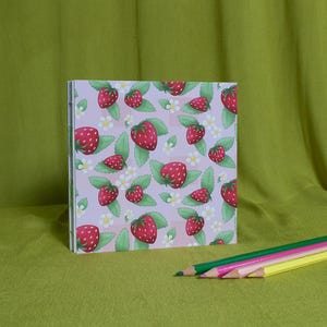 May include: A square notebook with a strawberry pattern on a pale purple background. The design features red strawberries, green leaves, and white flowers. Coloured pencils in green, pink, yellow, and light pink are in the foreground.