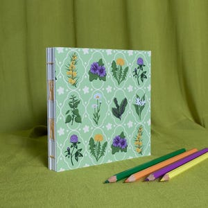 May include: A green journal with a floral design, showcasing diverse flowers and plants. The journal is bound with visible stitching and is next to four coloured pencils: green, orange, purple, and yellow. The background is a plain green fabric.