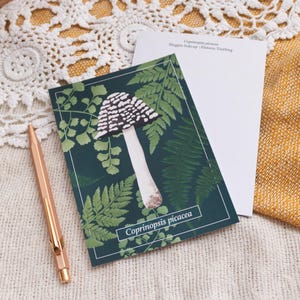 May include: A postcard with a mushroom illustration, ferns, and the text "Coprinopsis picaea." The card has a dark green background and white border. A blank card and a rose gold pen are also visible. The scene is set on a textured surface.