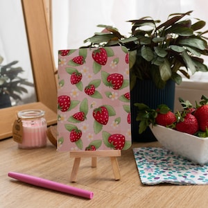 May include: A pink notebook with a strawberry pattern on the cover. The notebook is on a wooden easel and a pink pen is next to it.