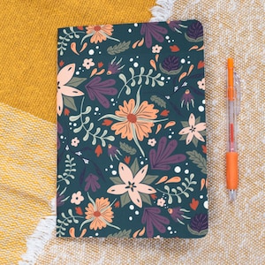 Op de afbeelding: Een donkergroen notitieboek met een bloemenpatroon in tinten roze, paars en wit. Het notitieboek ligt op een geel en wit textuur oppervlak.