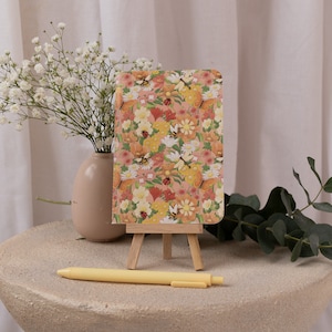 Op de afbeelding: Een klein notitieboekje met een bloemenpatroon op een houten ezel met een gele pen op een lichtgrijs oppervlak. Het notitieboekje heeft een bloemenpatroon met vlinders en lieveheersbeestjes.