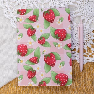 May include: A rectangular notebook with a light pink background featuring a repeating pattern of red strawberries, green leaves, and small white flowers. A solid light pink retractable ballpoint pen is positioned vertically next to the notebook.