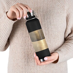 May include: Black insulated water bottle with silver accents. The bottle has three horizontal bands in gold, silver, and black. The bottle is held by a person.