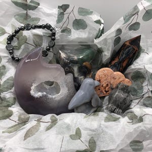 May include: A collection of crystals and stones in a white tissue paper gift bag. The crystals include a purple amethyst, a grey dolphin, a pink and white cloud, a black and grey stone, and a black hematite bracelet.