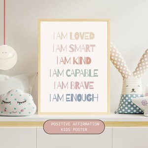 May include: A framed poster with a light wood frame and a white background. The poster features a positive affirmation for children with the text "I AM LOVED, I AM SMART, I AM KIND, I AM CAPABLE, I AM BRAVE, I AM ENOUGH". The text is in a variety of pastel colors. The poster also includes the text "POSITIVE AFFIRMATION KIDS POSTER" at the bottom.