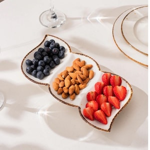 Könnte beinhalten: Eine weiße Keramikservierschale mit drei Abschnitten, die jeweils verschiedene Lebensmittel enthalten. Ein Abschnitt hat Blaubeeren, ein anderer Mandeln und der letzte Erdbeeren. Die Schale hat einen dekorativen braunen Rand.