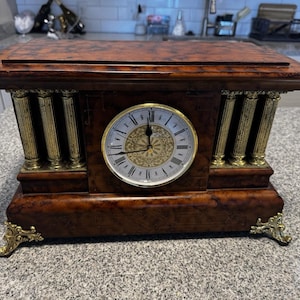May include: A vintage wooden mantel clock with a rectangular shape. The clock features a round face with Roman numerals and ornate gold detailing. Gold-coloured columns and decorative feet add to its classic design.