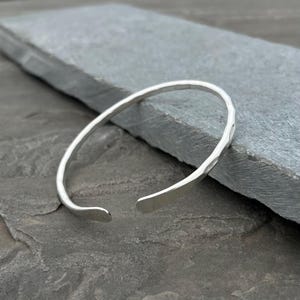 May include: A silver-colored open cuff bracelet with a hammered texture. The bracelet has a simple, elegant design and is open at the bottom. The metal has a bright, reflective finish, and the bracelet is displayed on a gray stone surface.