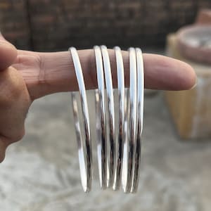 May include: A collection of silver bangles, each with a smooth, reflective surface. The bangles are displayed on a finger, showcasing their circular design and polished finish. The light reflects off the metal, creating a bright, eye-catching display.