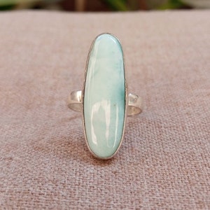 May include: A silver ring with a light blue oval-shaped gemstone. The ring is set on a light brown fabric background.
