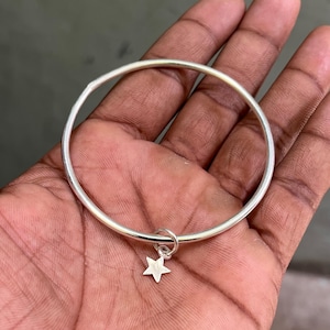 Könnte beinhalten: Ein silbernes Armreif-Armband mit einem kleinen Sternanhänger. Das Armband hat ein schlichtes, kreisförmiges Design mit glatter, polierter Oberfläche. Der Sternanhänger ist mit einer kleinen Schlaufe befestigt. Das Armband wird auf einer Hand gezeigt.