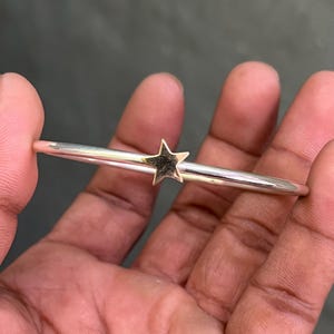 Puede incluir: Un brazalete plateado con un dije de estrella dorada. La estrella está centrada en el brazalete y tiene una superficie texturizada. El brazalete se sostiene en una mano. El diámetro es de unos 6 cm.