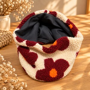 Könnte beinhalten: Ein runder Schlauchschal aus cremefarbenem Sherpa-Fleece mit einem wiederkehrenden Muster aus großen, dunkel kastanienbraunen Blumen mit leuchtend orangefarbenen Mitten. Die Innenseite ist mit einem einfarbigen, dunkel anthrazitgrauen Stoff gefüttert.