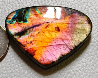 May include: A polished, teardrop-shaped labradorite gemstone with vibrant colors. The stone displays a mix of orange, pink, purple, and blue hues. A U.S. quarter is next to the gemstone for size comparison.