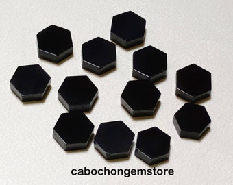 Può includere: Una collezione di dodici cabochon esagonali neri, probabilmente pietre preziose, disposti in cerchio su una superficie bianca. I cabochon sono lisci e lucidi, con una finitura lucida.