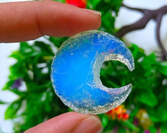 May include: A crescent moon shaped piece of blue opal with a white iridescent sheen. The opal is held in a hand against a background of green leaves and colorful flowers.