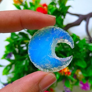 Pode incluir: Uma peça de opala azul em forma de lua crescente com um brilho iridescente branco. A opala é segurada na mão contra um fundo de folhas verdes e flores coloridas.