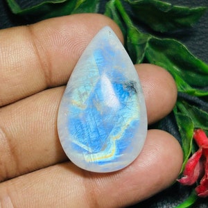 Peut inclure: Un cabochon de pierre de lune en forme de larme, iridescent, avec un jeu de couleurs bleu et blanc. La pierre a une surface lisse et polie et est sertie dans une bélière en argent.