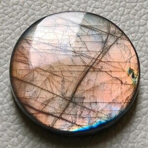 Peut inclure: Une pierre précieuse ronde et polie, aux couleurs variées, notamment rose, orange et bleu. La pierre présente un bord sombre et un réseau de fines lignes. Elle est posée sur un fond gris clair texturé.