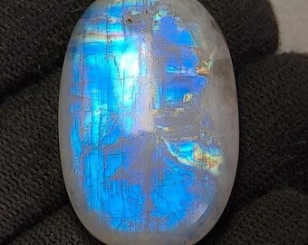 Può includere: Pietra preziosa di pietra di luna ovale con superficie lucida. La pietra mostra una vibrante adularescenza blu, con lampi di colore sulla sua superficie. La gemma è posta su uno sfondo scuro, evidenziando le sue qualità iridescenti.