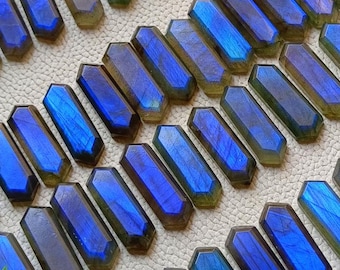 Könnte beinhalten: Eine Sammlung von 25 regenbogenfarbenen blauen und grünen Labradorit-Edelsteinen in facettierter, rechteckiger Form. Die Steine sind in Reihen auf einer weißen Oberfläche angeordnet.