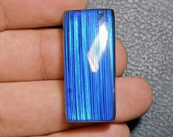 Puede incluir: Un cabujón de labradorita azul y blanco iridiscente con forma rectangular. La piedra preciosa tiene un patrón estriado y una superficie reflectante.