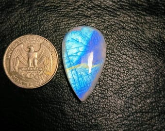 May include: A teardrop-shaped, polished gemstone with a vibrant blue and white color scheme, reminiscent of a celestial body. The gemstone is placed next to a U.S. quarter for size comparison. The stone has a cracked, web-like pattern.