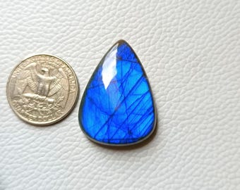 May include: A teardrop-shaped, polished blue gemstone with internal veining, next to a US quarter coin. The gemstone has a vibrant, iridescent quality, reflecting light. The coin shows the profile of George Washington. The background is a textured white surface.