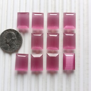 May include: A collection of twelve rectangular, translucent, pink gemstones. The gemstones are arranged in three rows of four, with a US quarter coin next to them for size comparison. The gemstones have a polished finish.