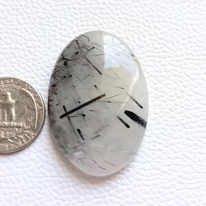 May include: Oval-shaped gemstone with clear and white tones, featuring black needle-like inclusions. A U.S. quarter coin is placed next to the gemstone for size comparison. The gemstone is set against a white textured surface.