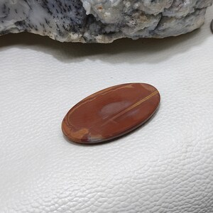 Puede incluir: Un cabuj&oacute;n liso de forma ovalada de jaspe rojo con finas vetas de color amarillo dorado. La piedra tiene un acabado pulido y un color natural y terroso.