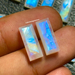 May include: Two iridescent rainbow moonstone cabochons, cut in a rectangular shape, with a white background and blue and yellow iridescent flashes. The stones are held in a hand.