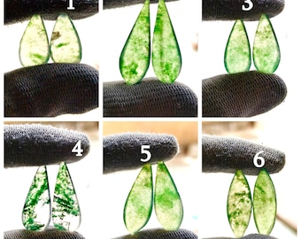 May include: Six pairs of teardrop-shaped green gemstones are displayed. Each pair is held between fingers, showcasing variations in color and pattern. The gemstones range from translucent to opaque, with inclusions creating unique designs. These gemstones are suitable for jewelry making.