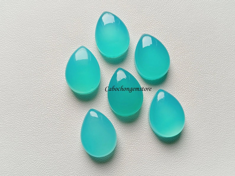 May include: Six teal blue teardrop shaped cabochon gemstones on a white surface.