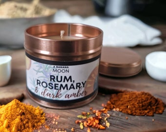 May include: A copper candle tin with a white label that reads "A Banana Moon, Rum, Rosemary & dark amber". The candle is sitting on a wooden surface with various spices around it.