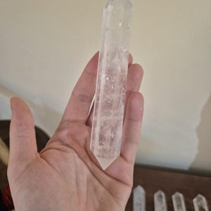 May include: A clear quartz crystal point, also known as a wand, is held in a hand. The crystal is long and slender with a pointed tip. The crystal is smooth and polished.