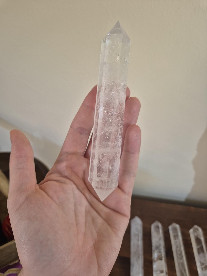 May include: A clear quartz crystal point, also known as a wand, is held in a hand. The crystal is long and slender with a pointed tip. The crystal is smooth and polished.