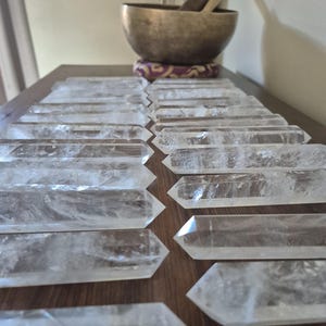 May include: A collection of clear quartz crystal points, also known as wands, arranged on a wooden surface. The crystals are smooth and polished, with a slight shimmer.
