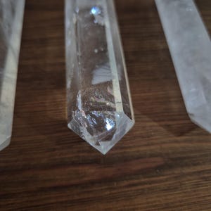 May include: Four clear quartz crystal points, also known as wands, are arranged on a wooden surface. The points are long and slender, with a pointed tip and a flat base.