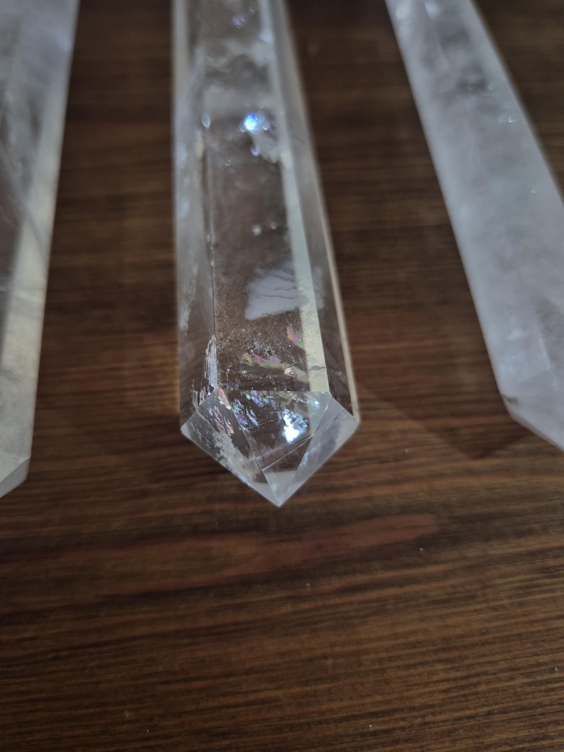 May include: Four clear quartz crystal points, also known as wands, are arranged on a wooden surface. The points are long and slender, with a pointed tip and a flat base.