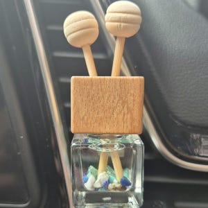 May include: A car air freshener with a wooden base and two wooden sticks. The glass bottle is filled with a clear liquid and small blue, white, and green pieces.