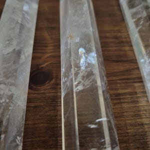 May include: A set of six clear quartz crystal points, each with a hexagonal shape and a pointed tip. The crystals are arranged in a row on a wooden surface.