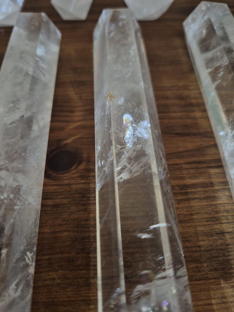 May include: A set of six clear quartz crystal points, each with a hexagonal shape and a pointed tip. The crystals are arranged in a row on a wooden surface.