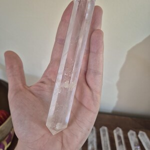 May include: A clear quartz crystal wand with a pointed tip. The wand is approximately 6 inches long and is held in a hand for size reference.