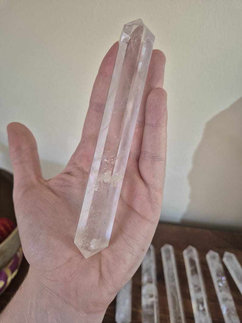 May include: A clear quartz crystal wand with a pointed tip. The wand is approximately 6 inches long and is held in a hand for size reference.