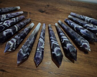 May include: A set of 12 amethyst wands with a white and purple striped pattern. The wands are pointed at both ends and are arranged in a circle on a wooden surface.
