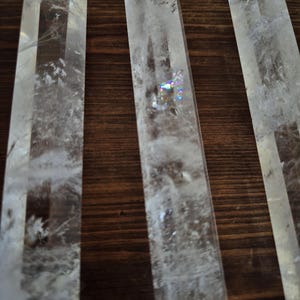 May include: Three clear quartz crystal points, also known as wands, are arranged side by side on a wooden surface. The crystals are long and slender, with a pointed tip and a flat base. They are all approximately the same size and shape.