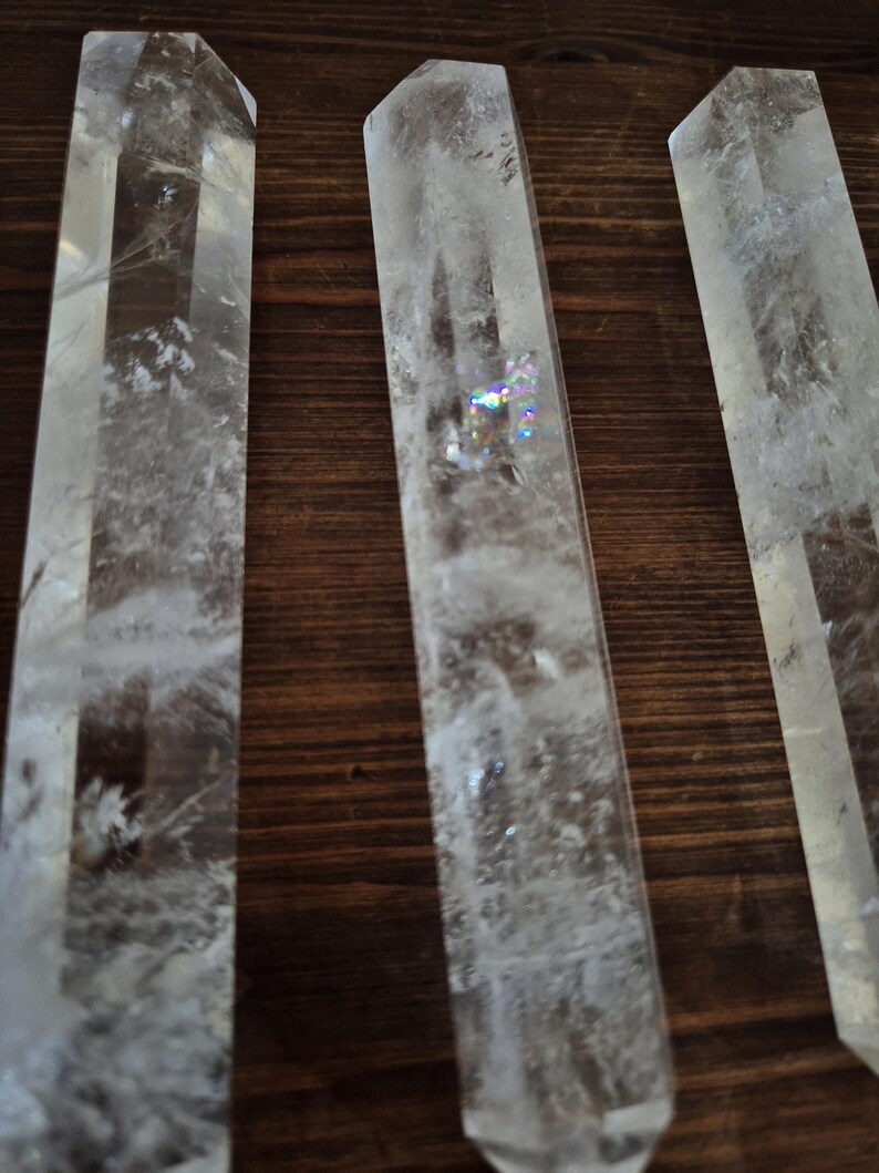 May include: Three clear quartz crystal points, also known as wands, are arranged side by side on a wooden surface. The crystals are long and slender, with a pointed tip and a flat base. They are all approximately the same size and shape.
